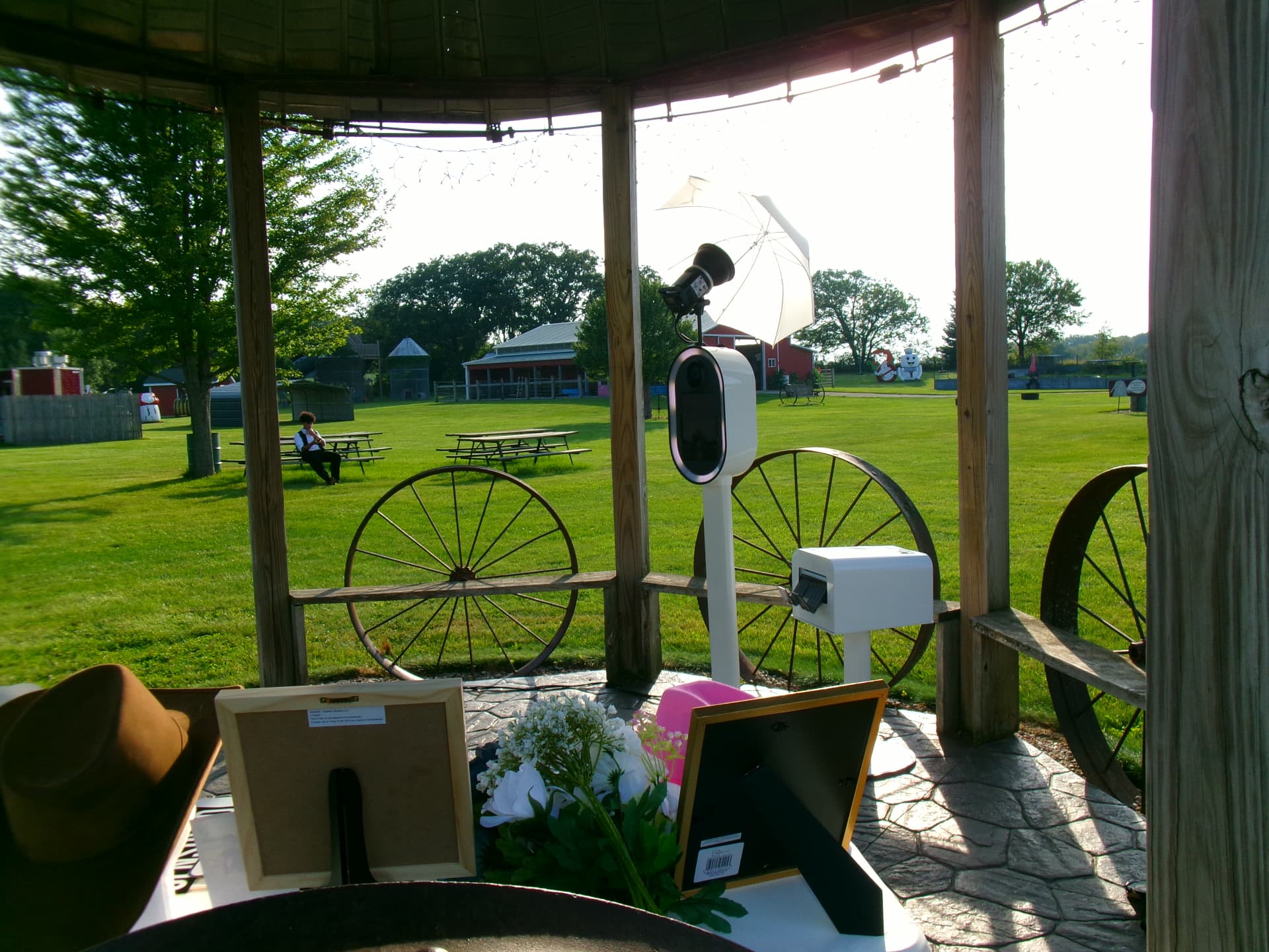 Memento photo booth and printer station set up inside a rustic wooden gazebo with string lights, overlooking a sunny lawn and barn at an outdoor Wisconsin venue